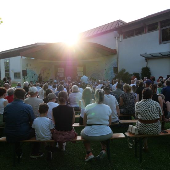 Eine Menschenmenge sitzt auf Bänken vor einem Gebäude und sieht sich bei Sonnenuntergang eine Aufführung auf der Bühne an.