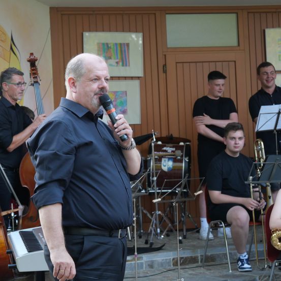 Ein Mann steht vor einem Mikrofon und spricht eine Gruppe von Musikern an, die sitzen und stehen und verschiedene Instrumente spielen. Die Musiker umfassen einen Cellisten, einen Schlagzeuger und einen Trompeter.