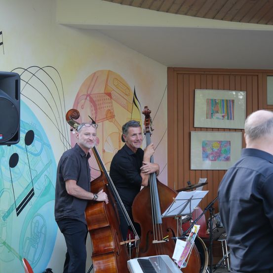 Zwei Männer spielen Kontrabass vor einer Wand mit Noten und Bildern.
