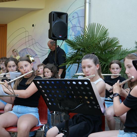 Eine Gruppe junger Mädchen spielt Flöten in einem Raum mit einer musikalischen Wand im Hintergrund. Ein Mann steht mit einem Lautsprecher hinter ihnen.