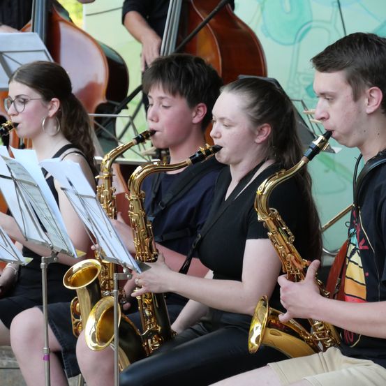 Vier Personen sitzen und spielen Saxophon, mit Notenblättern vor ihnen. Im Hintergrund befinden sich ein Cello und eine grüne Wand.