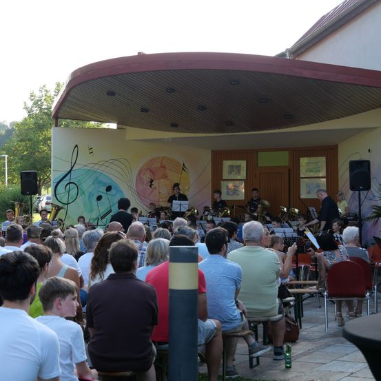 Ein Blasorchester spielt auf einer Bühne mit gemalten Musiknoten, das Publikum sitzt davor.