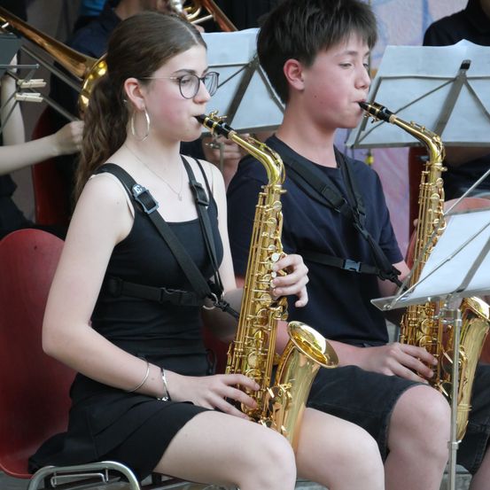 Zwei junge Musiker, ein Mädchen und ein Junge, spielen Saxophon in einer Band. Sie sitzen und tragen schwarze Gurte um ihre Schultern. Hinter ihnen spielen andere Bandmitglieder verschiedene Instrumente.