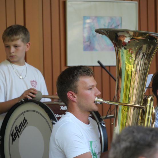 Ein junger Mann spielt Tuba, während ein anderer Junge hinter ihm mit einem Drumstick steht. Sie befinden sich in einem Raum mit Holzwänden.