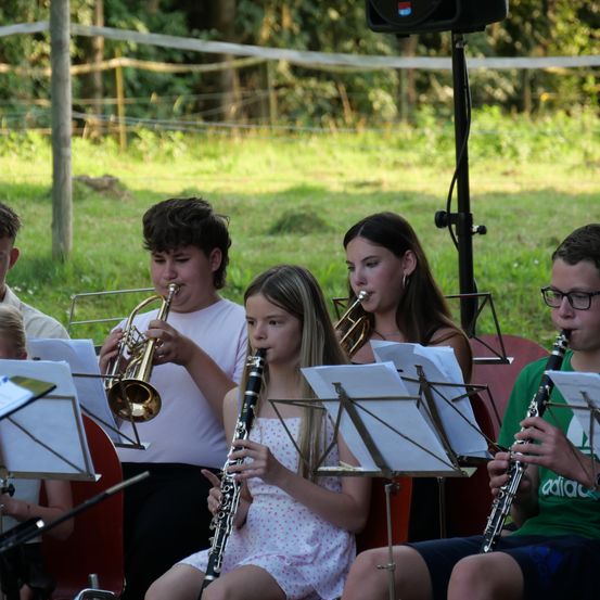 Eine Gruppe junger Musiker spielt Instrumente im Freien. Ein Junge spielt Trompete, ein Mädchen Klarinette und ein weiteres Mädchen Trompete. Sie sitzen auf Stühlen mit Notenständern vor ihnen.