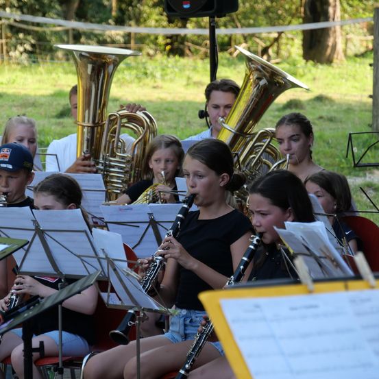 Eine Gruppe junger Musiker, darunter Mädchen und Jungen, übt im Freien mit verschiedenen Instrumenten wie Tubas, Trompeten und Klarinetten. Sie sitzen auf Stühlen, lesen Noten und spielen in einem grün bewachsenen Bereich.