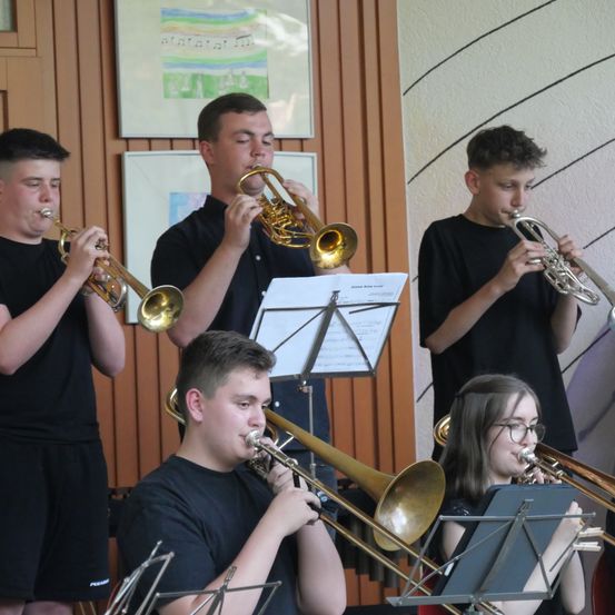 Fünf junge Musiker, in Schwarz gekleidet, spielen Trompeten und Posaunen in einem Raum mit Holzwänden und einer weißen Wand mit geometrischem Muster.