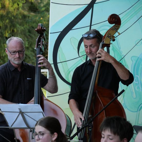 Zwei Männer spielen Cellos in einer Außenumgebung, mit einem musikalischen Noten-Hintergrund und einem jungen Mädchen und Jungen, die zusehen.