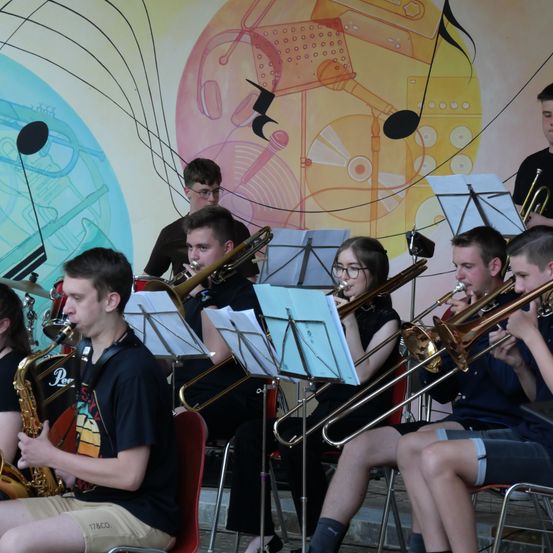 Eine Gruppe junger Musiker spielt auf der Bühne Instrumente, mit einem farbenfrohen Wandbild im Hintergrund.