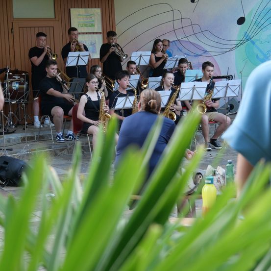 Eine Gruppe junger Musiker spielt auf der Bühne mit Trompeten und Saxophonen, mit einer farbenfrohen Wand im Hintergrund.