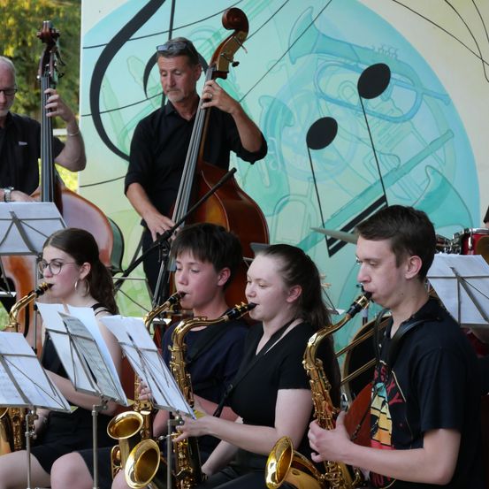 Eine Gruppe junger Musiker spielt Saxophon in einer Freiluftumgebung. Dahinter spielt ein Mann Cello und ein anderer Mann Kontrabass. Sie stehen vor einem Hintergrund mit Musiknoten und Instrumenten.