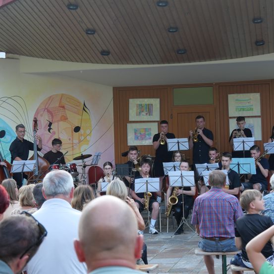 Eine Gruppe von Musikern spielt in einem Halbkreis auf einer Bühne mit einem farbenfrohen Wandbild im Hintergrund. Das Publikum sitzt vor ihnen.