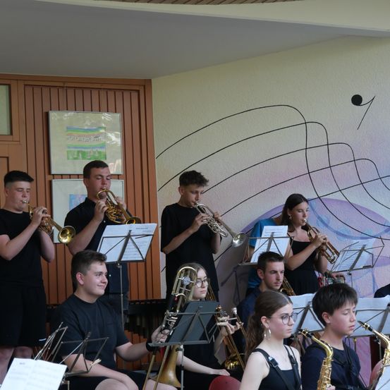 Eine Gruppe junger Musiker, sowohl Männer als auch Frauen, spielen Trompeten, Posaunen und Saxophone in einem Raum mit einem Wandbild an der Wand.
