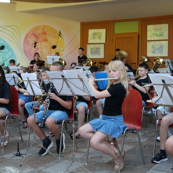 Eine Gruppe von Kindern in einem Musikraum, die verschiedene Instrumente wie Trompeten, Posaunen und Flöten spielen. Sie sitzen auf Stühlen mit Notenständern vor ihnen.