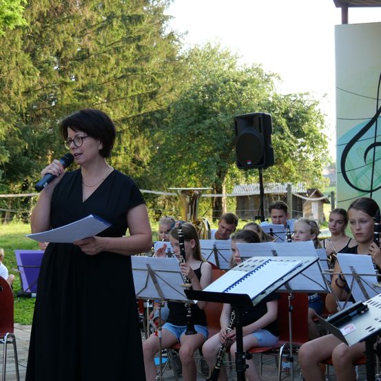 Eine Frau in einem schwarzen Kleid spricht in ein Mikrofon, während eine Gruppe von Kindern auf Stühlen sitzt und Instrumente spielt.