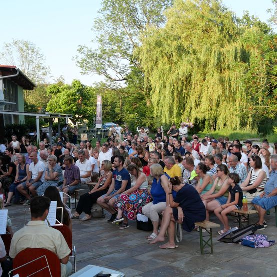 Eine große Menschenmenge versammelt sich im Freien, wahrscheinlich für ein Konzert oder eine Aufführung, auf Bänken und Stühlen unter Bäumen sitzend.