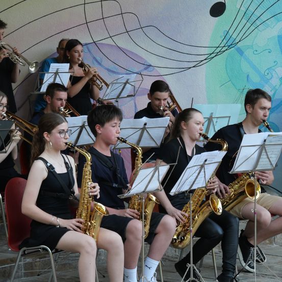 Eine Gruppe junger Musiker spielt Instrumente, darunter Saxophone, Trompeten und Posaunen, vor einer Wand mit Noten.