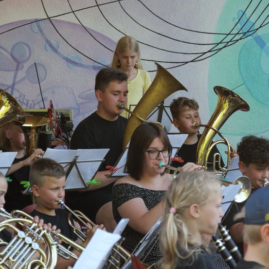 Eine Gruppe junger Musiker, darunter Jungen und Mädchen, spielt Blechblasinstrumente im Freien. Sie stehen vor einer Wand mit bunten musikalischen Illustrationen.