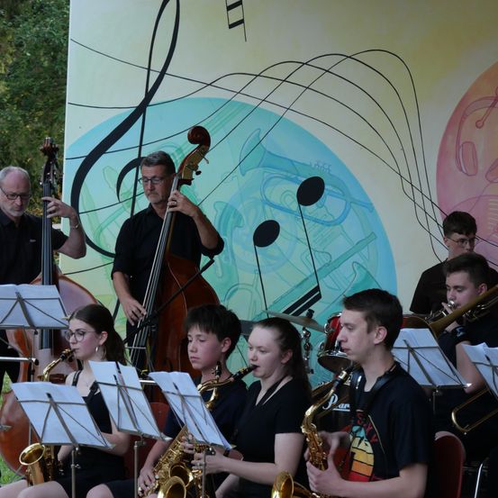 Eine Gruppe junger Musiker, darunter Saxophonisten, Trompeter und ein Bassist, spielen auf der Bühne vor einem Hintergrund mit Noten.