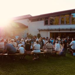 Eine große Gruppe von Menschen versammelt sich im Freien, sitzt auf Bänken und hört einem Redner zu. Die Sonne geht im Hintergrund unter und taucht die Szene in ein warmes Licht.