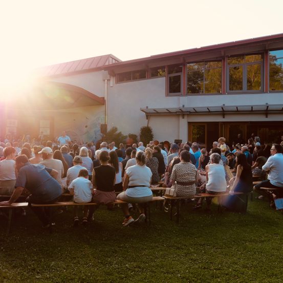 Eine große Gruppe von Menschen versammelt sich im Freien, sitzt auf Bänken und hört einem Redner zu. Die Sonne geht im Hintergrund unter und taucht die Szene in ein warmes Licht.