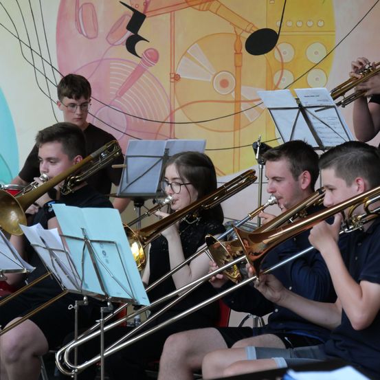 Eine Gruppe junger Musiker spielt Posaunen in einem Raum mit einer farbenfrohen Wand im Hintergrund.