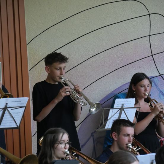 Eine Gruppe junger Musiker, darunter ein Junge und ein Mädchen, spielen Trompeten und Posaunen in einem Raum mit gemusterter Wand. Sie schauen auf ihre Notenblätter.