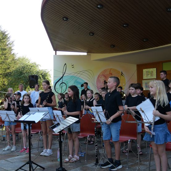 Eine Gruppe von Kindern in schwarzen Shirts und Shorts steht vor Musikständern mit Musikinstrumenten, wahrscheinlich in Vorbereitung auf eine Aufführung. Dahinter befindet sich eine Wand mit Noten und einem Wandbild.