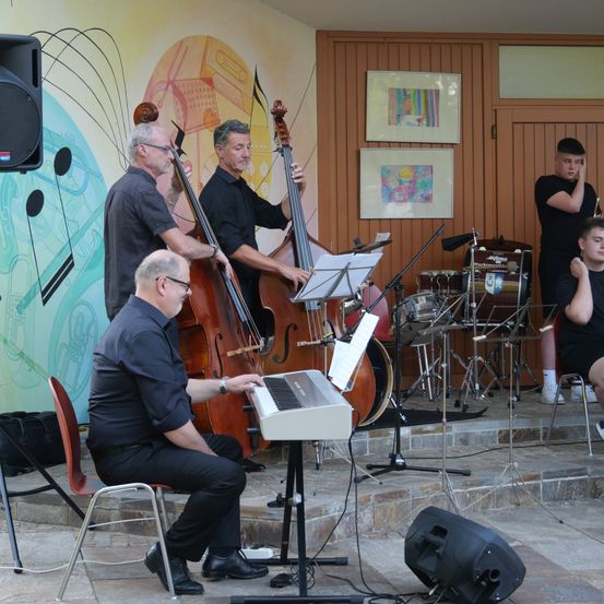Eine Gruppe von Musikern spielt auf einer Bühne mit Musikinstrumenten. Ein Mann spielt das Klavier, während ein anderer Mann das Cello spielt. Es gibt auch Trommeln und andere Instrumente auf der Bühne.