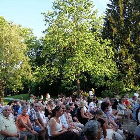 Eine Gruppe von Menschen sitzt in einem Halbkreis unter einem Blätterdach, wahrscheinlich bei einer Veranstaltung im Freien. Die Umgebung ist ein Park mit üppiger Grünfläche und klarem blauem Himmel.