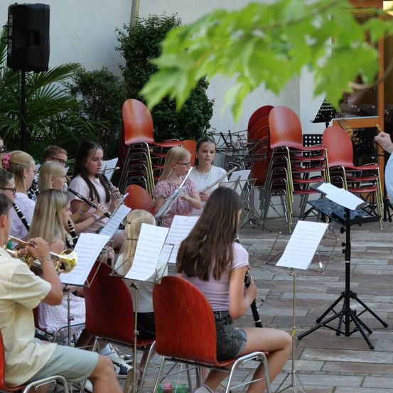 Eine Gruppe junger Musiker, darunter Mädchen und Jungen, spielt verschiedene Instrumente wie Trompeten, Flöten und Klarinetten auf einer Terrasse. Sie sitzen auf roten Stühlen mit Notenständern vor ihnen.