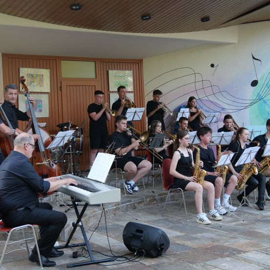 Eine Gruppe von Musikern spielt in einem Raum mit Noten an der Wand, einige sitzen und andere stehen und spielen verschiedene Instrumente.