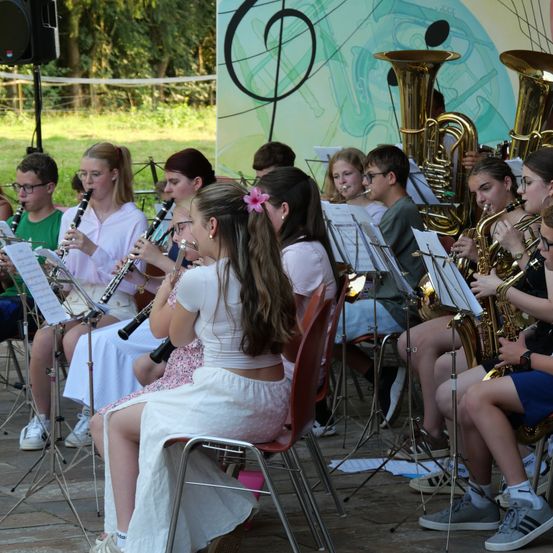 Eine Gruppe junger Musiker, darunter Mädchen und Jungen, sitzen auf Stühlen und spielen verschiedene Blasinstrumente wie Klarinetten und Trompeten in einer Außenumgebung.
