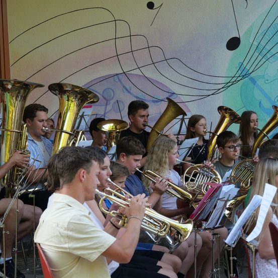 Eine Gruppe junger Musiker, sowohl Jungen als auch Mädchen, spielen verschiedene Blechblasinstrumente wie Trompeten, Posaunen und Tuben in einem Raum mit einem farbenfrohen musikalischen Hintergrund.