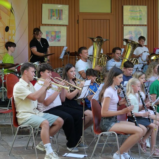 Eine Gruppe junger Musiker spielt auf der Bühne, mit Trompeten, Tubas und Klarinetten. Sie sitzen auf Stühlen, und eine Frau steht hinter ihnen, möglicherweise ihre Lehrerin.