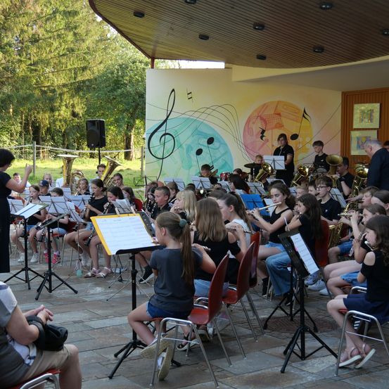 Eine Schulband probt im Freien mit einem Dirigenten, umgeben von Musikinstrumenten und einem farbenfrohen Wandbild.