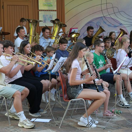 Eine Gruppe junger Musiker, sowohl Jungen als auch Mädchen, spielen verschiedene Instrumente im Freien. Sie sitzen auf Stühlen und stehen, halten Trompeten, Posaunen und andere Blechblasinstrumente. Sie tragen Freizeitkleidung und scheinen sich in einer Probensitzung zu befinden.