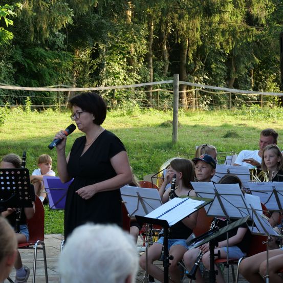 Eine Frau mit Brille steht vor einer Gruppe von Kindern, die in einem Grasfeld Instrumente spielen.