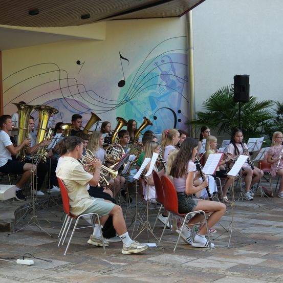 Eine Gruppe von Musikern, sowohl jung als auch erwachsen, sitzen auf Stühlen und spielen verschiedene Instrumente, darunter Trompeten, Posaunen und Klarinetten, vor einer Wand mit Noten und einem Lautsprecher.