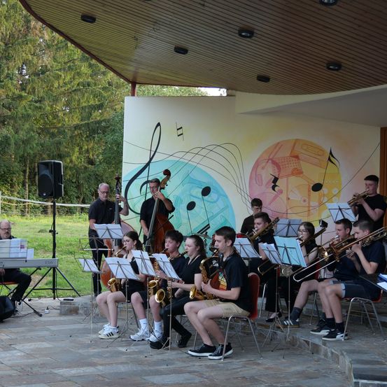 Eine Gruppe von Musikern spielt auf einer Bühne mit musikalischen Noten, die an der Wand hinter ihnen gemalt sind. Einige sitzen, während andere stehen.
