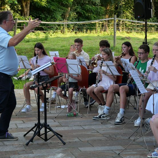 Eine Gruppe junger Musiker, darunter Mädchen und Jungen, spielen verschiedene Instrumente wie Trompeten und Klarinetten unter der Anleitung eines Mannes in einem blauen Hemd. Sie sitzen auf Stühlen in einer Außenumgebung mit einem Grasbereich und einem Zaun im Hintergrund.