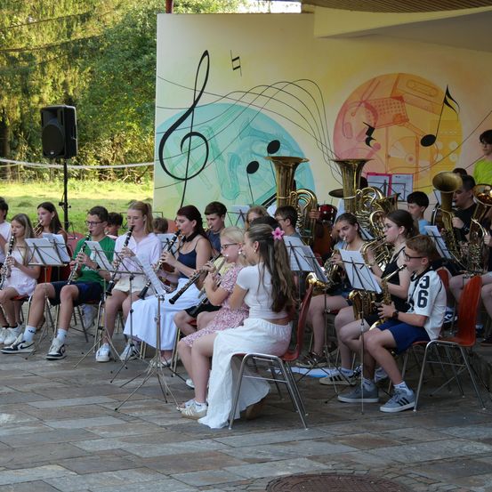 Eine Gruppe junger Musiker spielt auf der Bühne mit verschiedenen Instrumenten, darunter Klarinetten und Trompeten, vor einer Wand mit gemalten Musiknoten und Instrumenten.