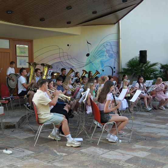 Eine Gruppe junger Musiker, darunter Trompeten- und Klarinettenspieler, sitzen auf Stühlen und spielen auf einer Bühne mit Noten, die an der Wand hinter ihnen gemalt sind.