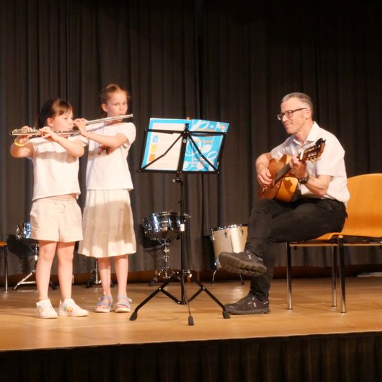 Zwei junge Mädchen spielen Flöten, während ein Mann Gitarre auf einer Bühne spielt, mit einem Notenständer vor ihnen.