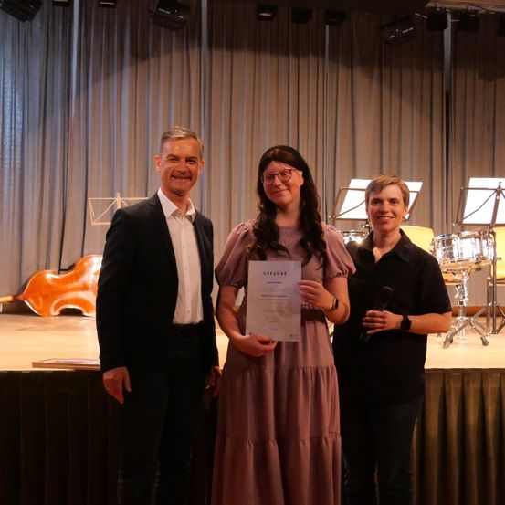 Drei Personen stehen auf einer Bühne, eine hält ein Zertifikat. Eine Frau in der Mitte lächelt und hält das Zertifikat, während zwei Männer neben ihr stehen. Hinter ihnen sind Musikinstrumente und ein Vorhang zu sehen.