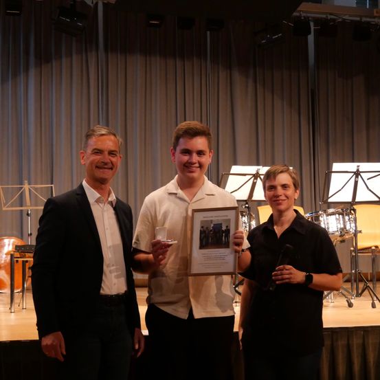 Drei Personen stehen auf einer Bühne, zwei halten ein gerahmtes Zertifikat und einen Glas, lächeln. Dahinter sind Musikinstrumente auf Ständern aufgestellt.