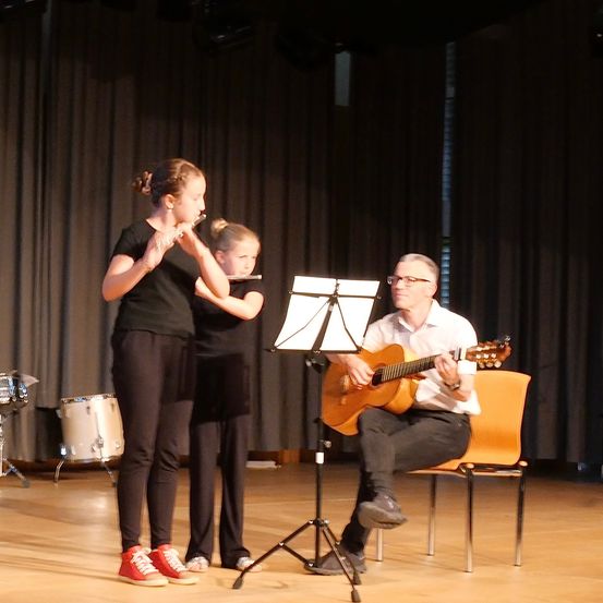 Zwei Mädchen und ein Mann treten auf der Bühne auf. Ein Mädchen spielt Flöte, das andere Flöte und der Mann Gitarre.