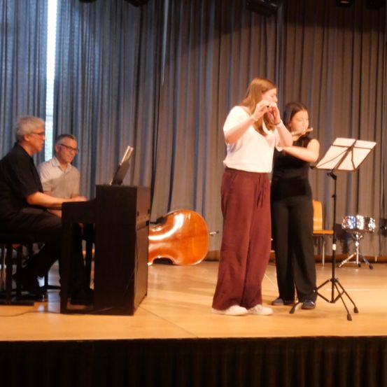 Zwei Frauen spielen Flöten auf der Bühne, während ein Mann im Hintergrund Klavier spielt.