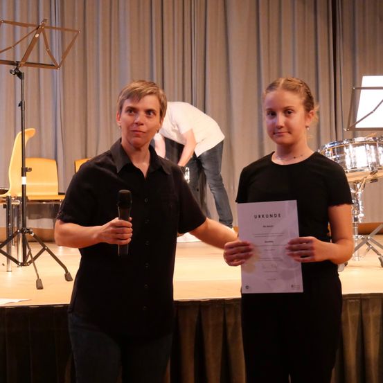 Eine Frau und ein Mädchen stehen auf einer Bühne, die Frau hält ein Mikrofon und das Mädchen ein Zertifikat. Hinter ihnen ist ein Mann, der etwas auf der Bühne justiert.