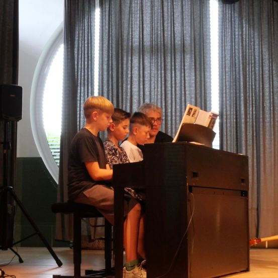 Drei Jungen und eine ältere Frau sitzen an einem Klavier, wobei die Frau Noten liest. Die Jungen scheinen sich auf die Klaviertasten zu konzentrieren.